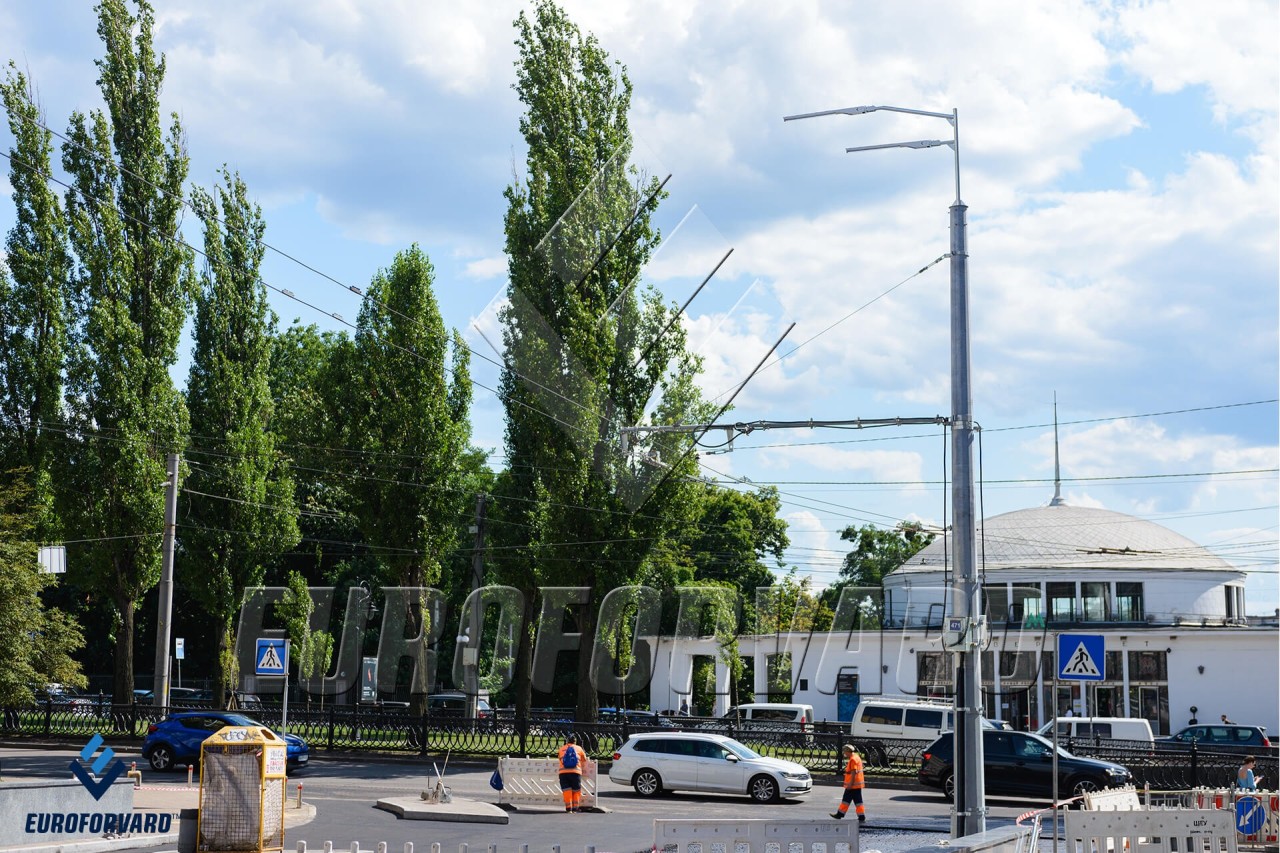 Опори контактної мережі біля дороги на фоні будівлі метро